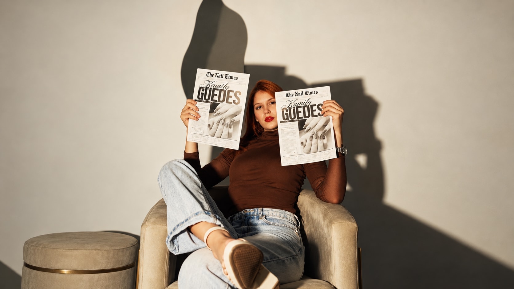 Kamily Guedes sentada em uma poltrona ao sol, segurando duas edições do jornal The Nail Times com seu nome na capa